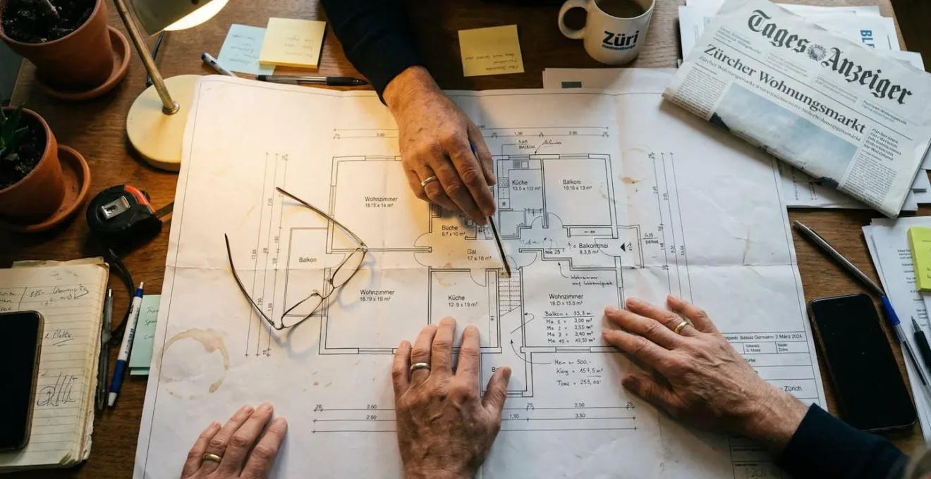 Hände zeigen auf einen Grundriss auf Papier, Kugelschreiber und Brille auf dem Tisch, Schweizer Zeitung im Hintergrund